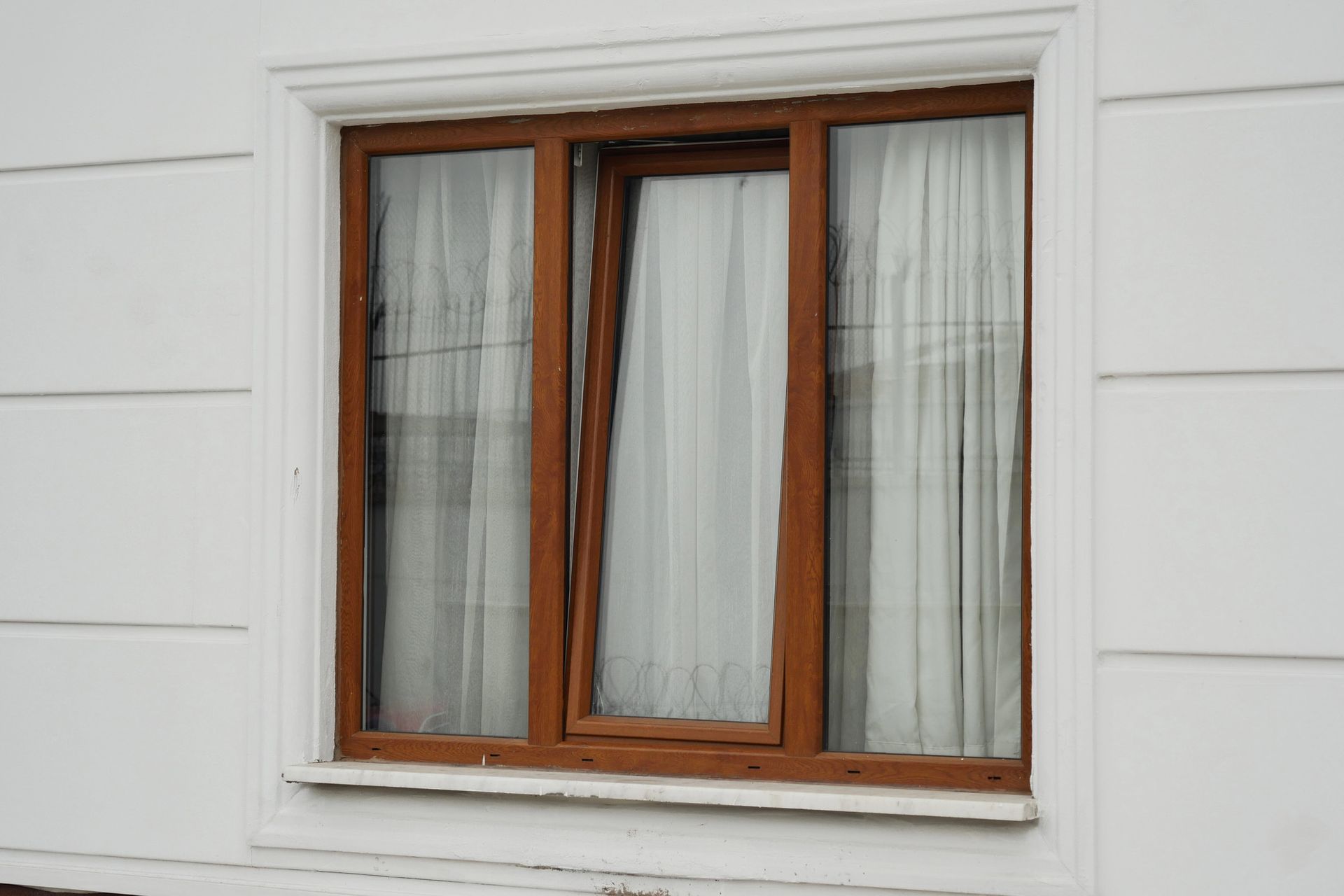 Parete bianca con finestra con cornice in legno marrone e tende trasparenti.