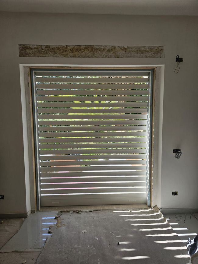 Vista interna di una porta da garage con lamelle orizzontali; la luce del sole proietta ombre a strisce sul pavimento.