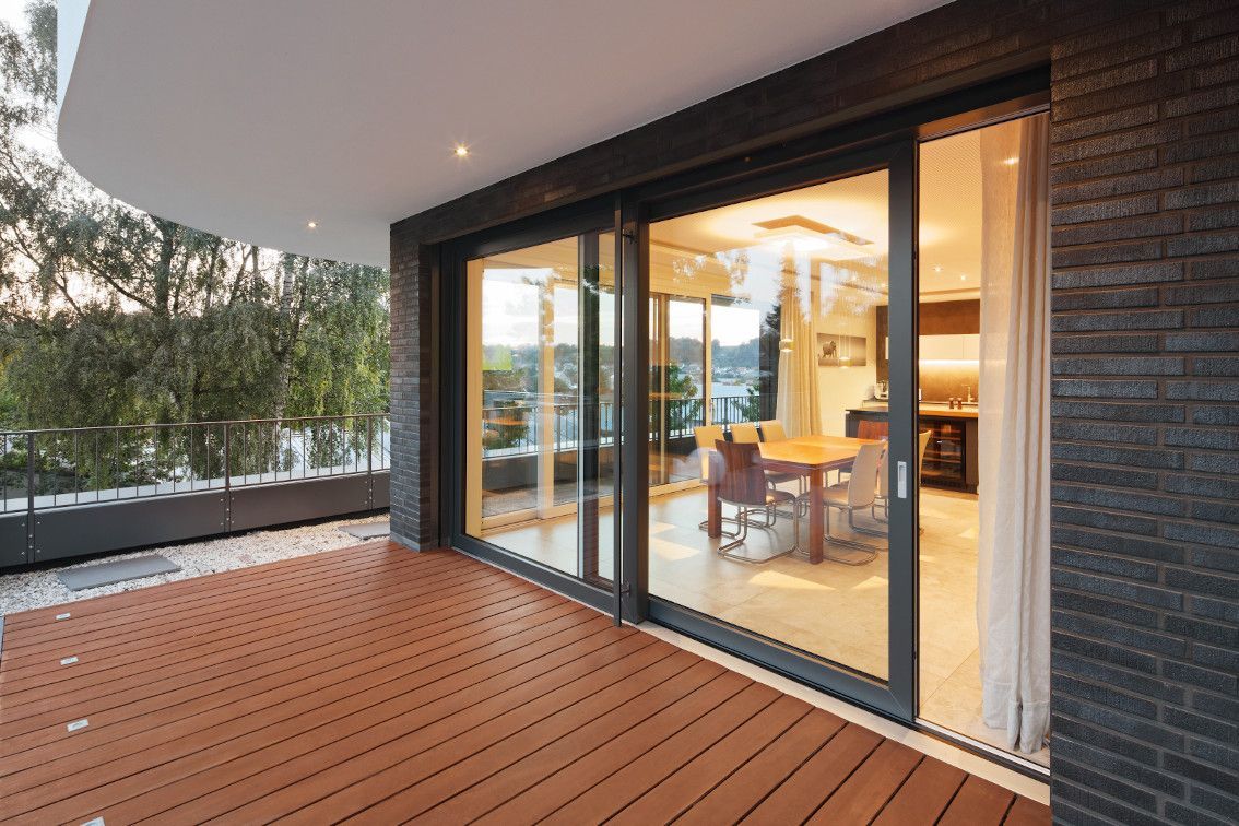 Patio moderno con pavimentazione in legno, esterno in mattoni e porte scorrevoli in vetro che conducono a un interno luminoso.