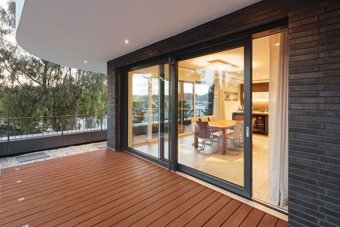 Moderno balcone con pavimentazione in legno e porte scorrevoli in vetro che si aprono su un interno illuminato da una luce calda.