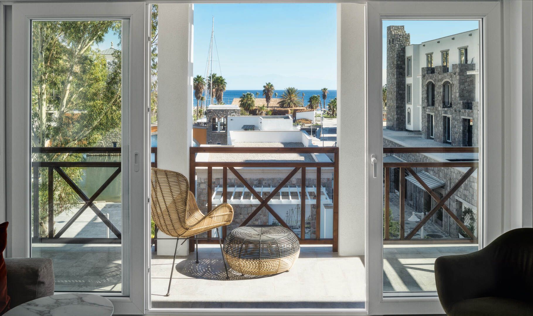 Vista dal balcone su un porticciolo turistico sul mare, con sedia e tavolo in vimini incorniciati da porte aperte.