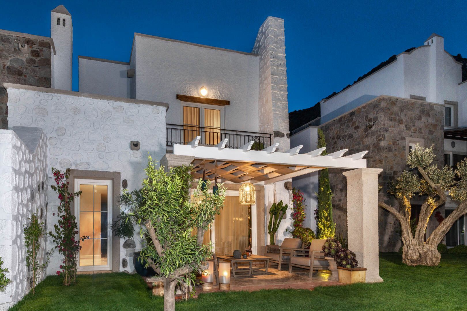 Casa moderna in stucco bianco con luci da patio, pergola e prato curato al crepuscolo