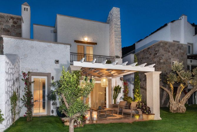 Casa moderna in stucco bianco con luci da patio, pergola e prato curato al crepuscolo