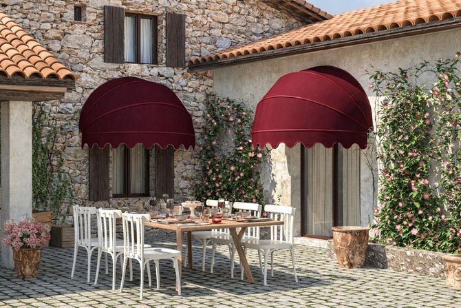 Zona pranzo con patio in pietra, sedie bianche, tavolo in legno e tende da sole bordeaux sulla facciata rustica di una villa.