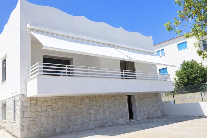 Casa moderna bianca a due piani con seminterrato in pietra, balcone e cielo azzurro brillante.
