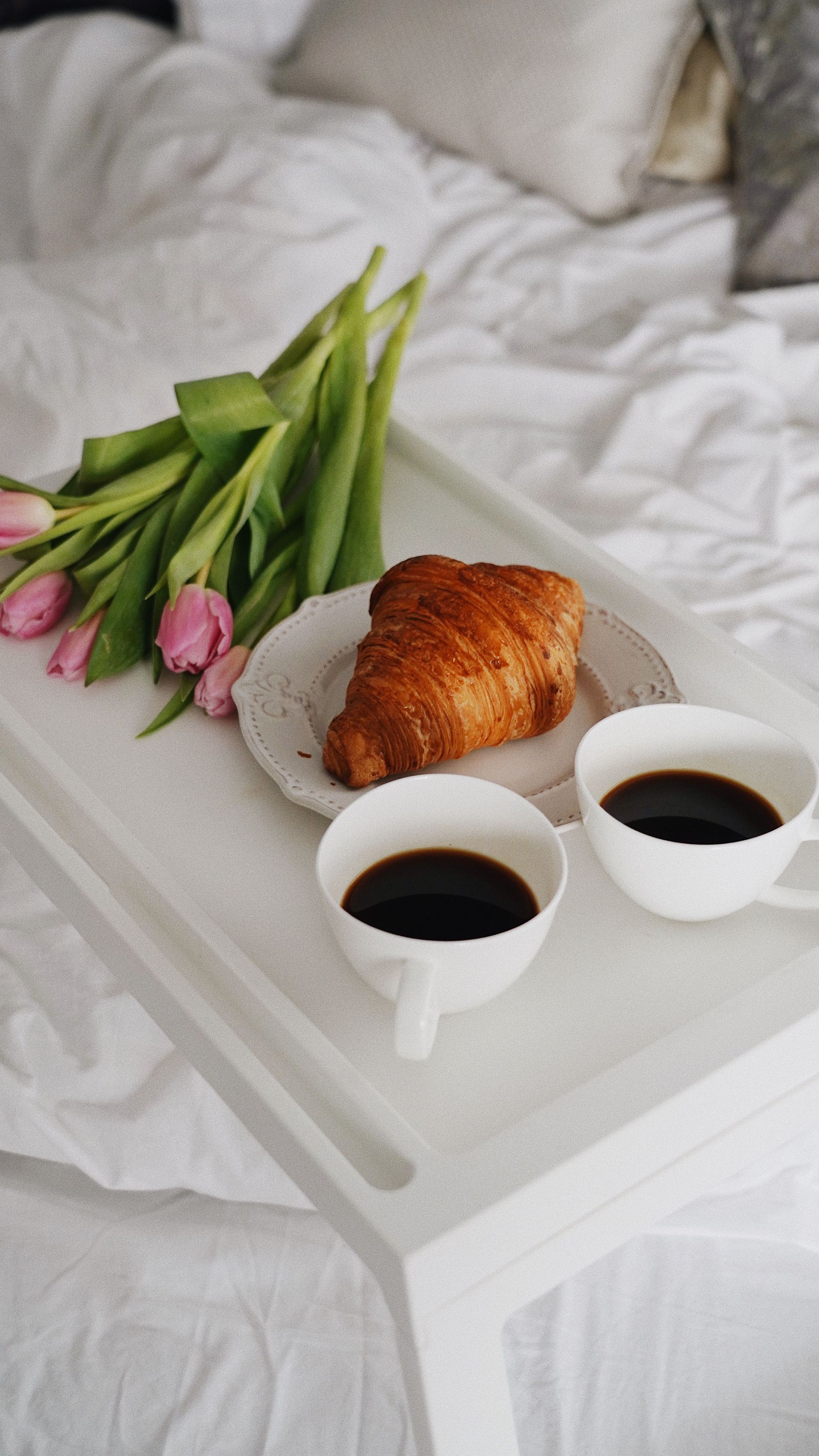 Two cups of black coffee beside a croissant and pink tulips — illustrative photo