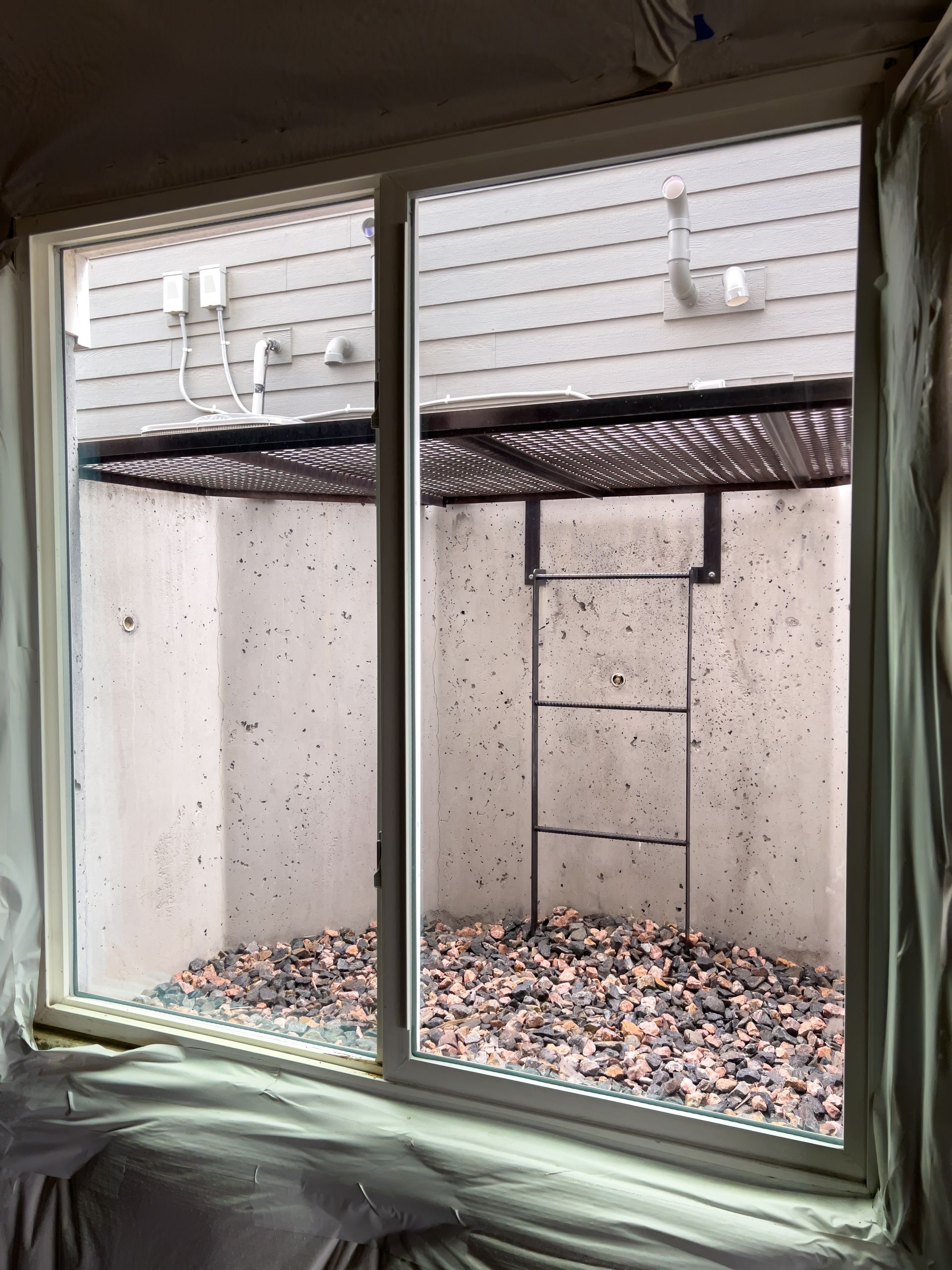 White-framed sliding window with view of a sunken window well filled with gravel and a ladder. White-framed sliding window with view of a sunken window well filled with gravel and a ladder.