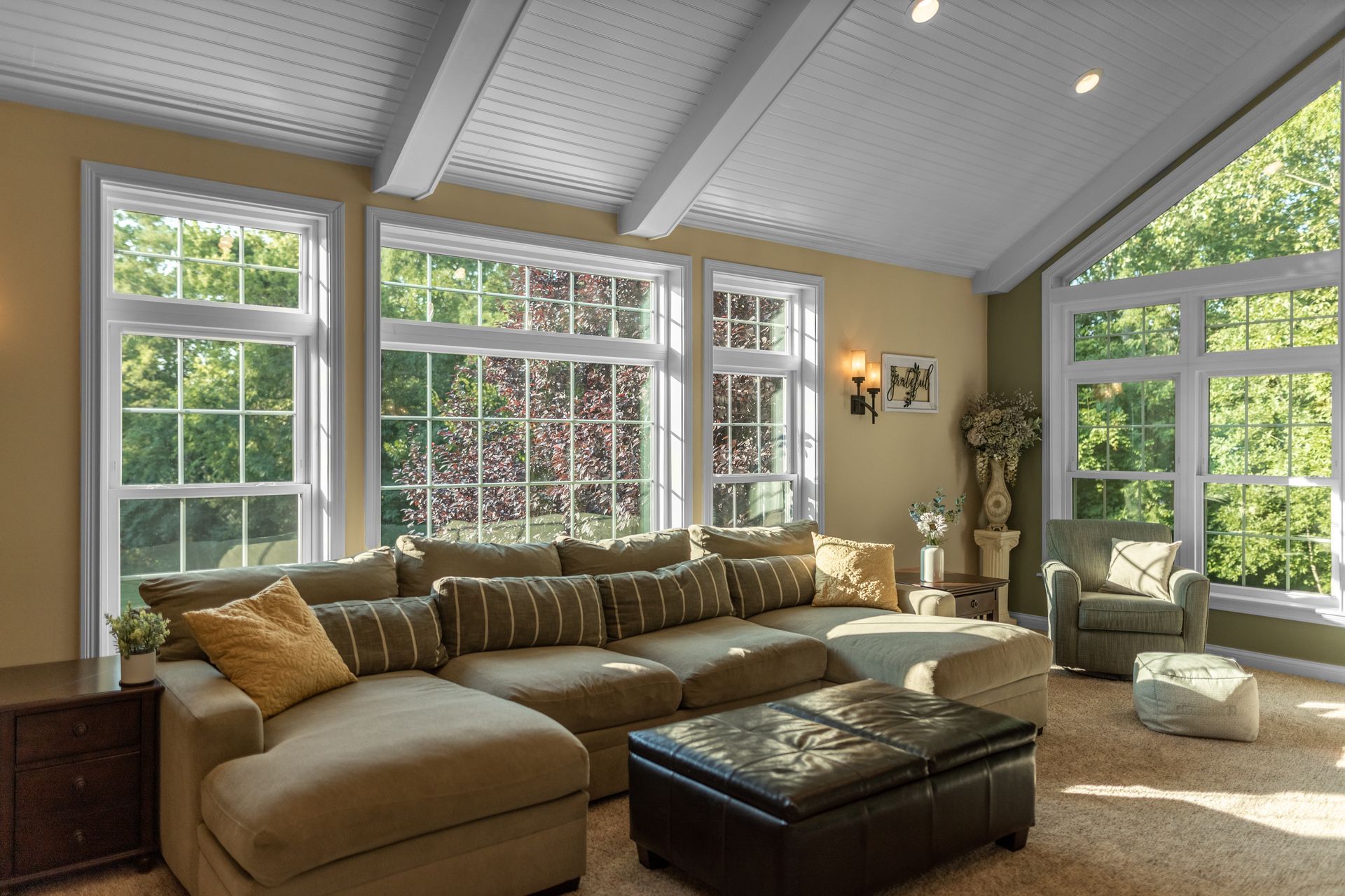 Living room with large windows, sectional sofa, and ottoman. Beige walls and white ceiling. Living room with large windows, sectional sofa, and ottoman. Beige walls and white ceiling.