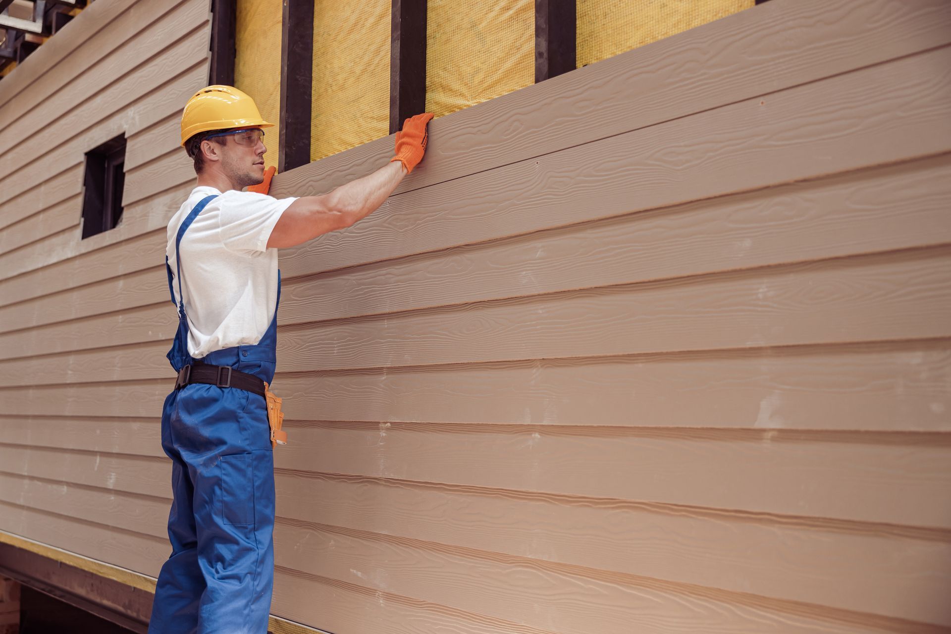 Construction worker in overalls, hard hat, and gloves installing beige siding on a building's exterior.