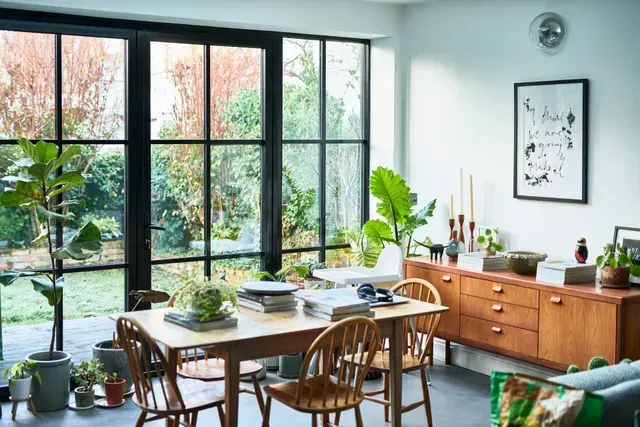 Dining room with large window overlooking garden, table, chairs, plants, and wooden sideboard.