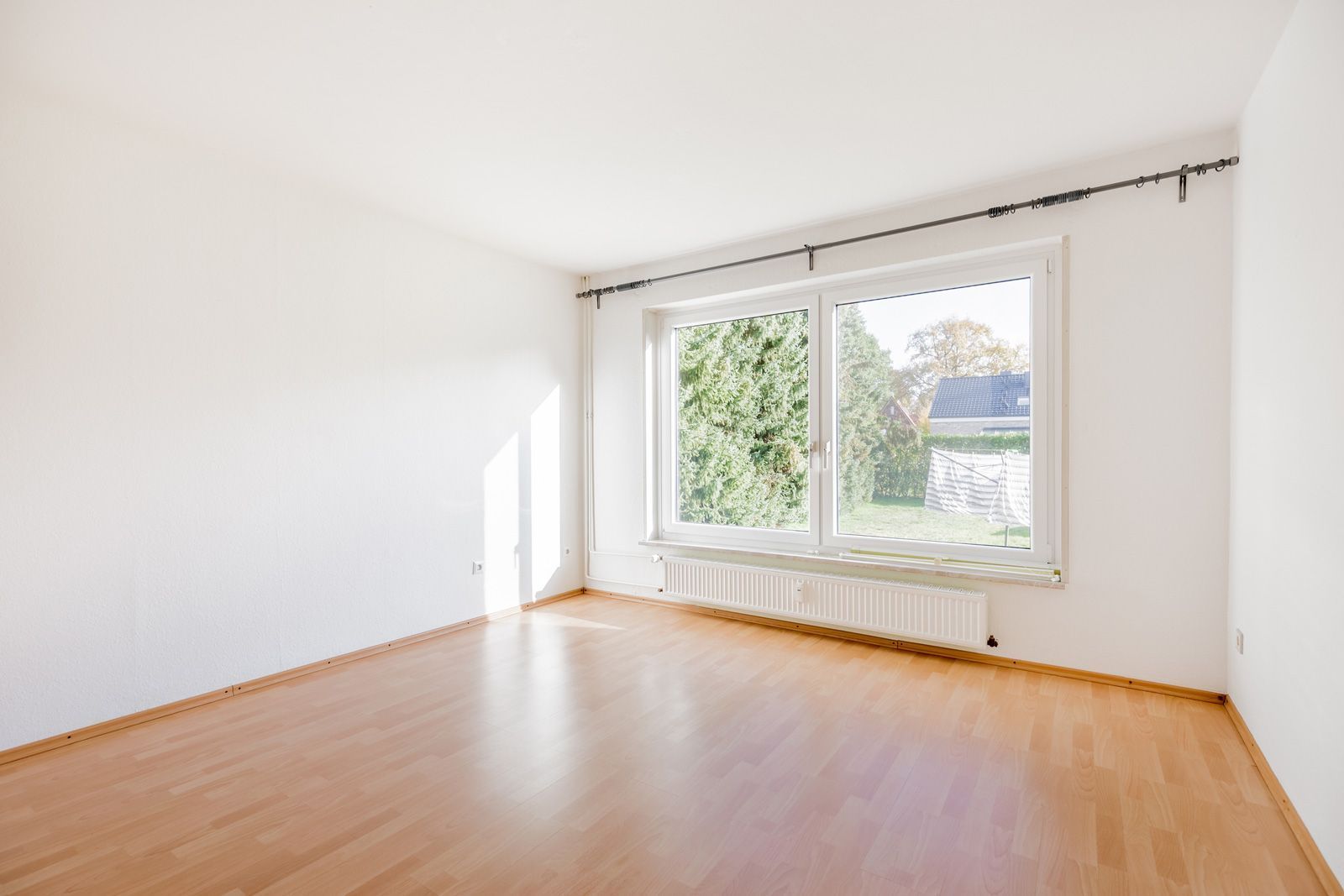 Empty room with wooden floor, white walls, and large window overlooking trees.