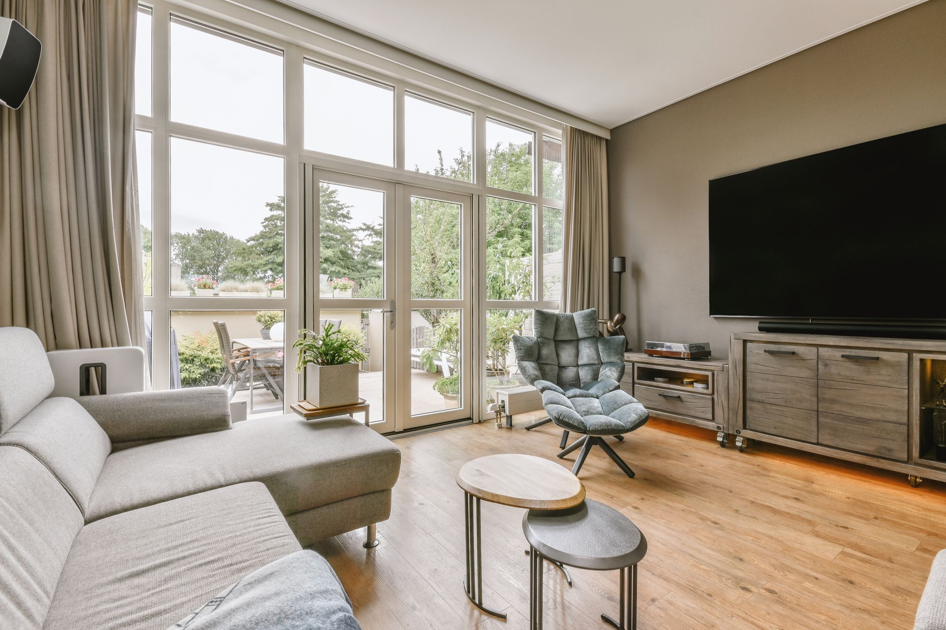 Living room with gray sofa, large TV, windows, and small round coffee tables. Living room with gray sofa, large TV, windows, and small round coffee tables.