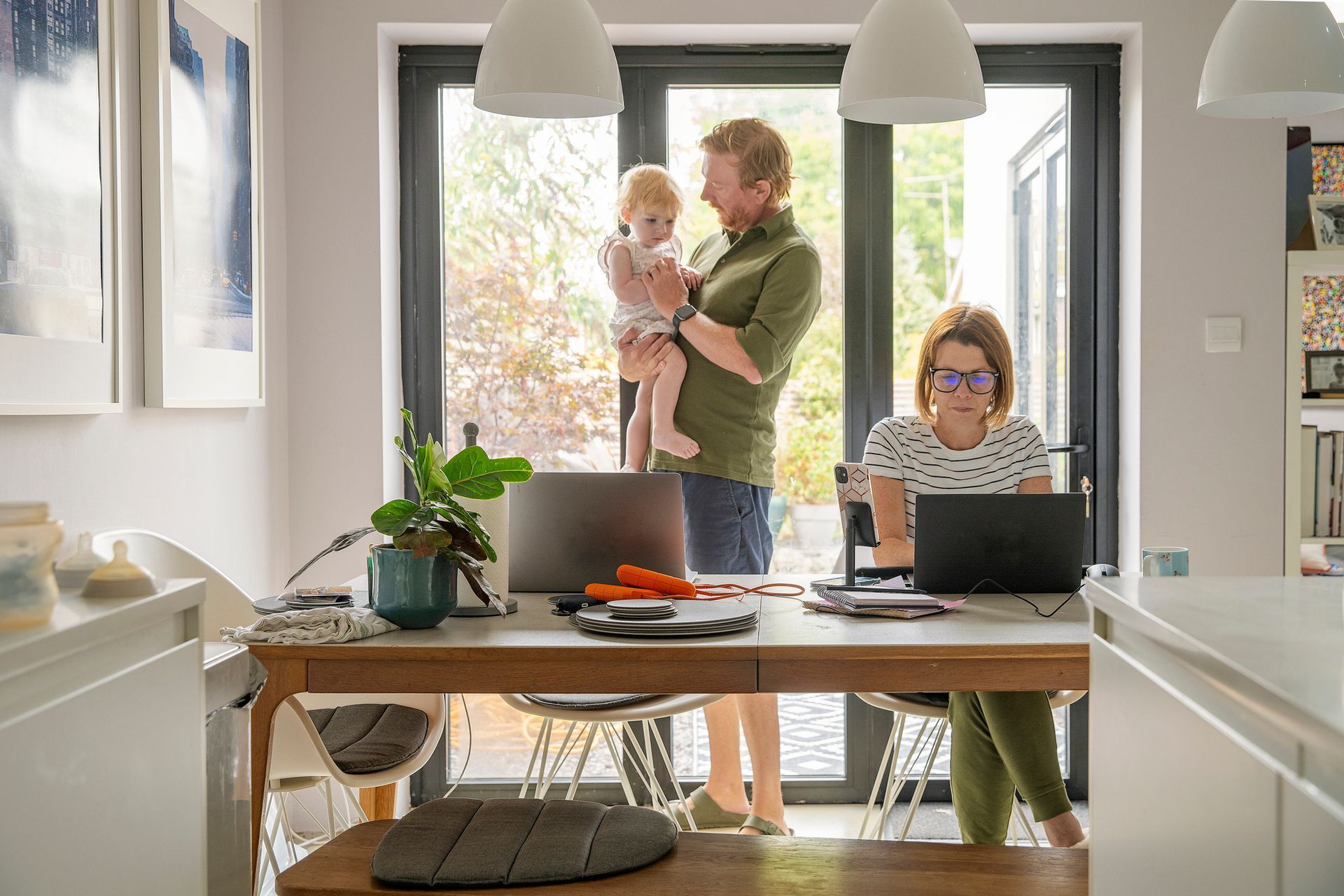 Family working at home: parent holding child, another working on a laptop, sunny room with open doors. Family working at home: parent holding child, another working on a laptop, sunny room with open doors.