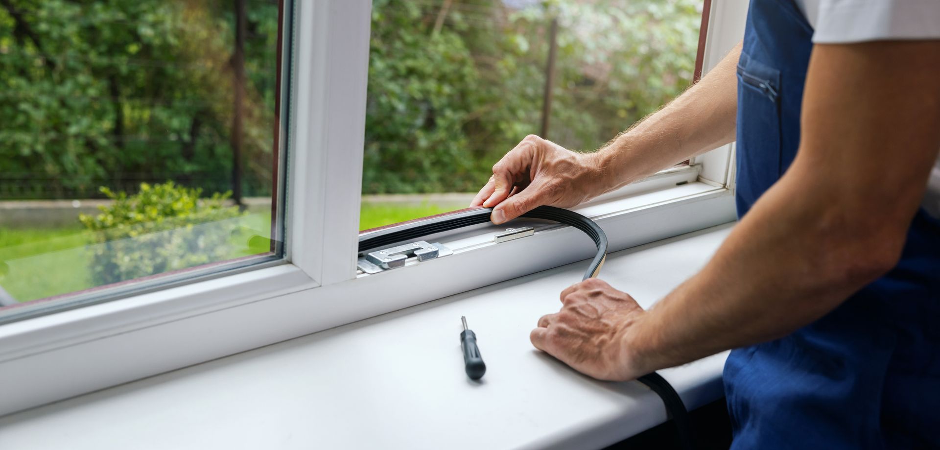 Person installing weather stripping on a white window frame, holding a strip, with a screwdriver nearby. Person installing weather stripping on a white window frame, holding a strip, with a screwdriver nearby.