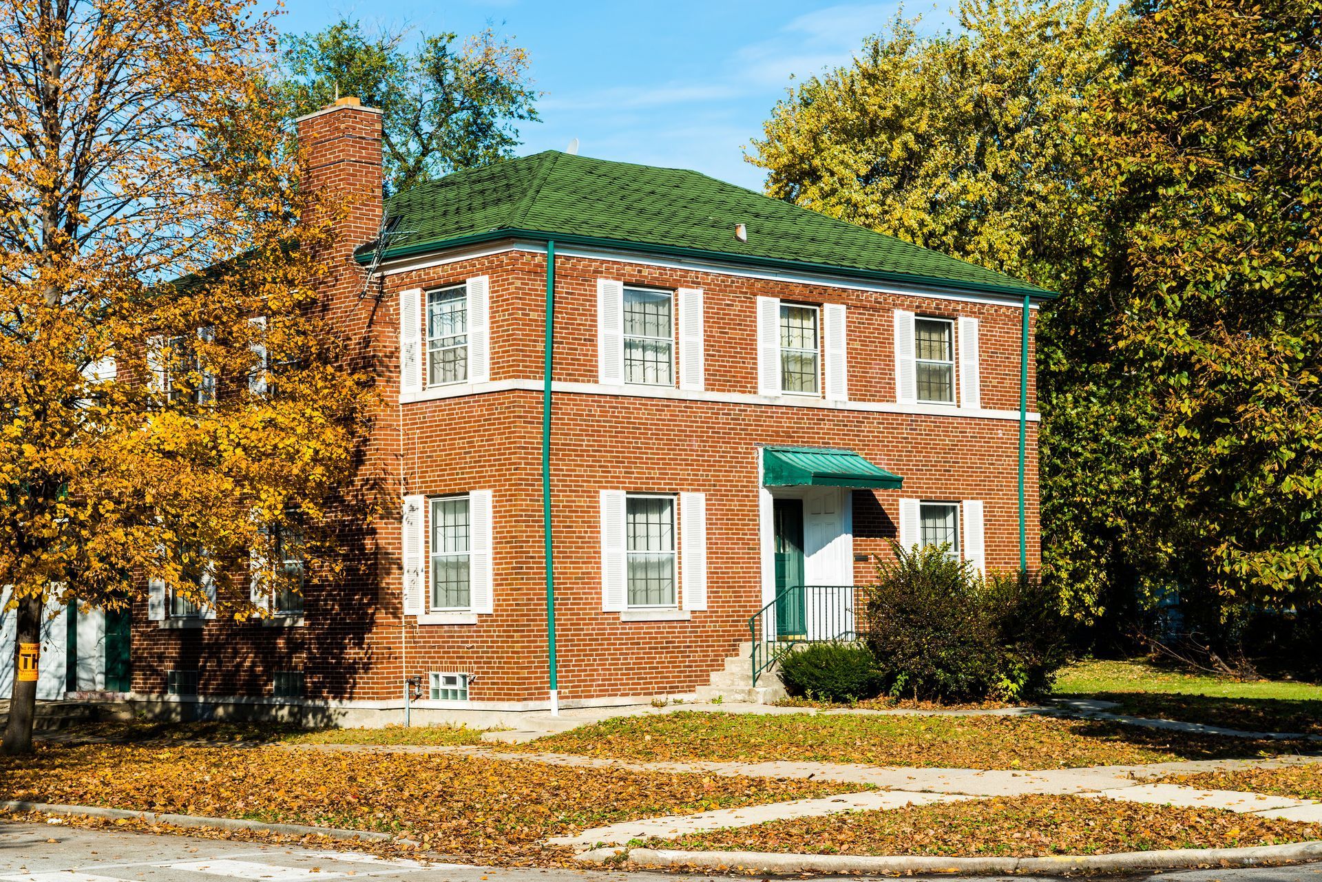 Two-story brick house with green roof, white shutters, and fall foliage.