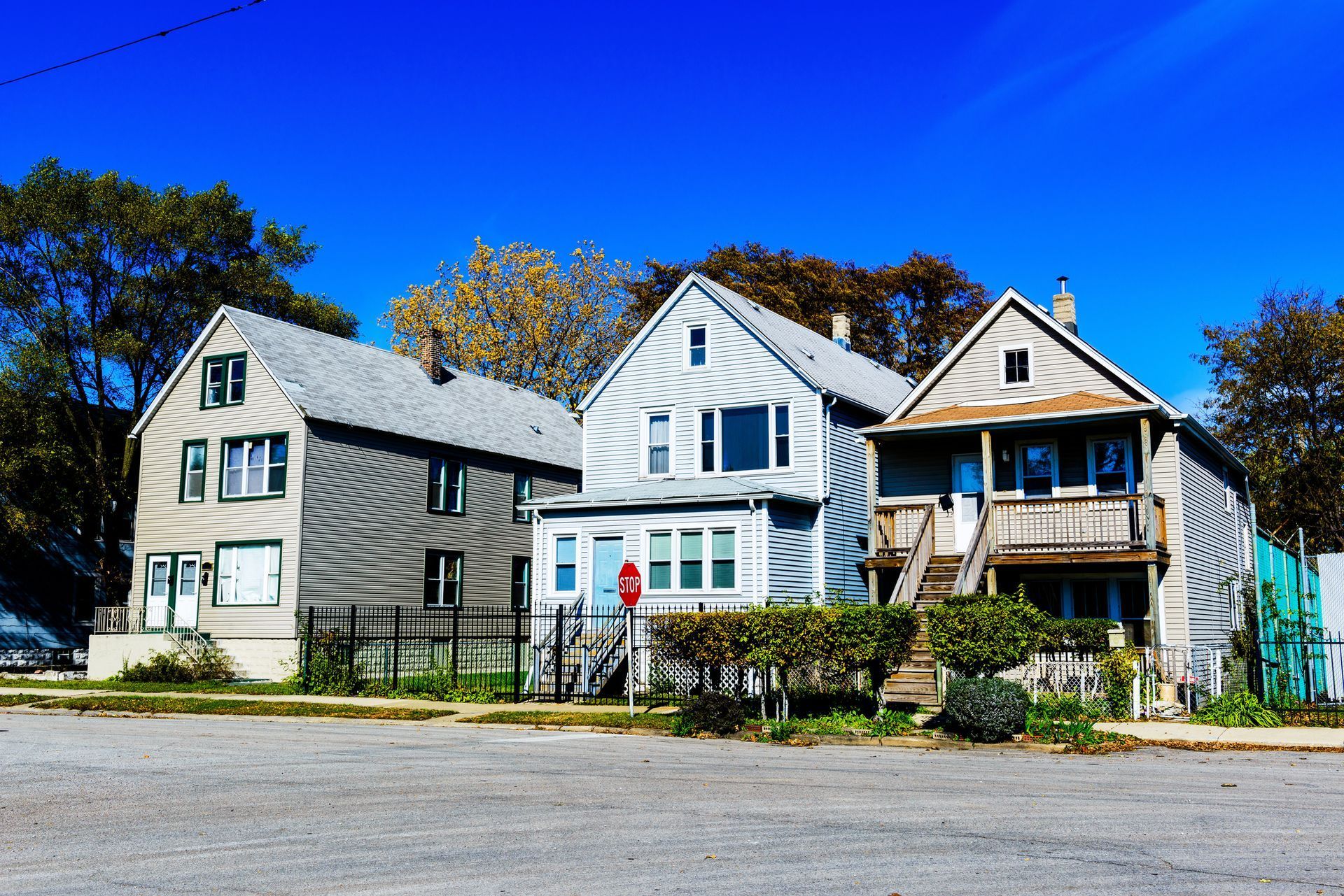 Row of three houses, varying colors, against a clear blue sky. Two-story buildings with pitched roofs and visible front doors.