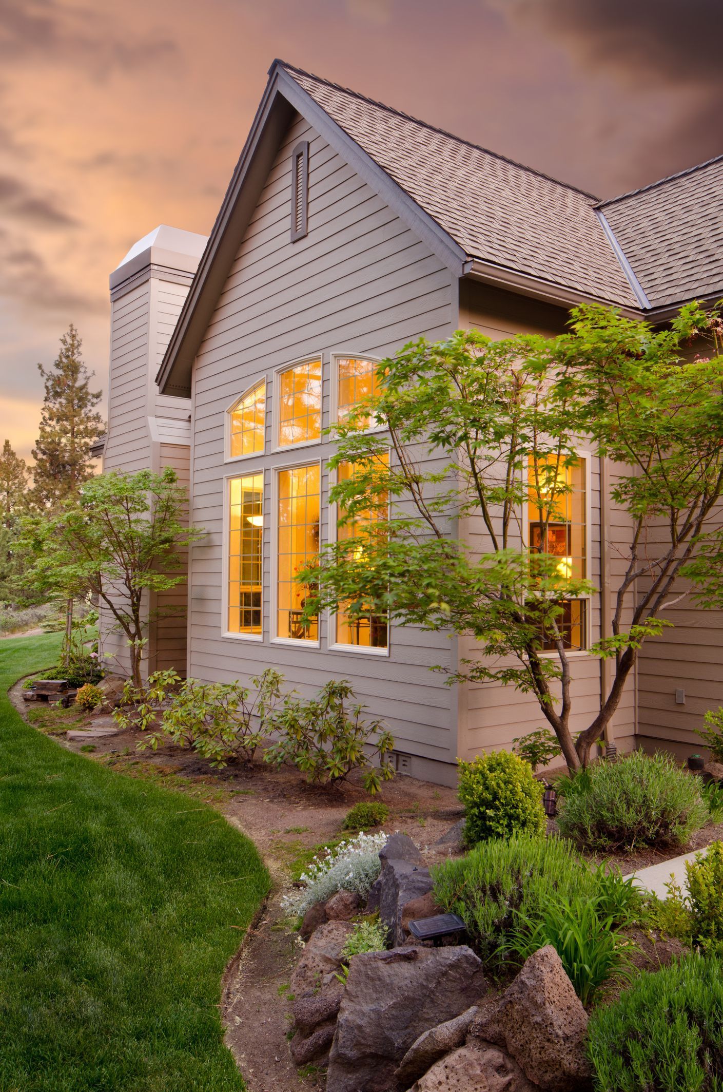 Exterior of a house with a stone garden and large windows lit from within, under a sunset sky. Exterior of a house with a stone garden and large windows lit from within, under a sunset sky.
