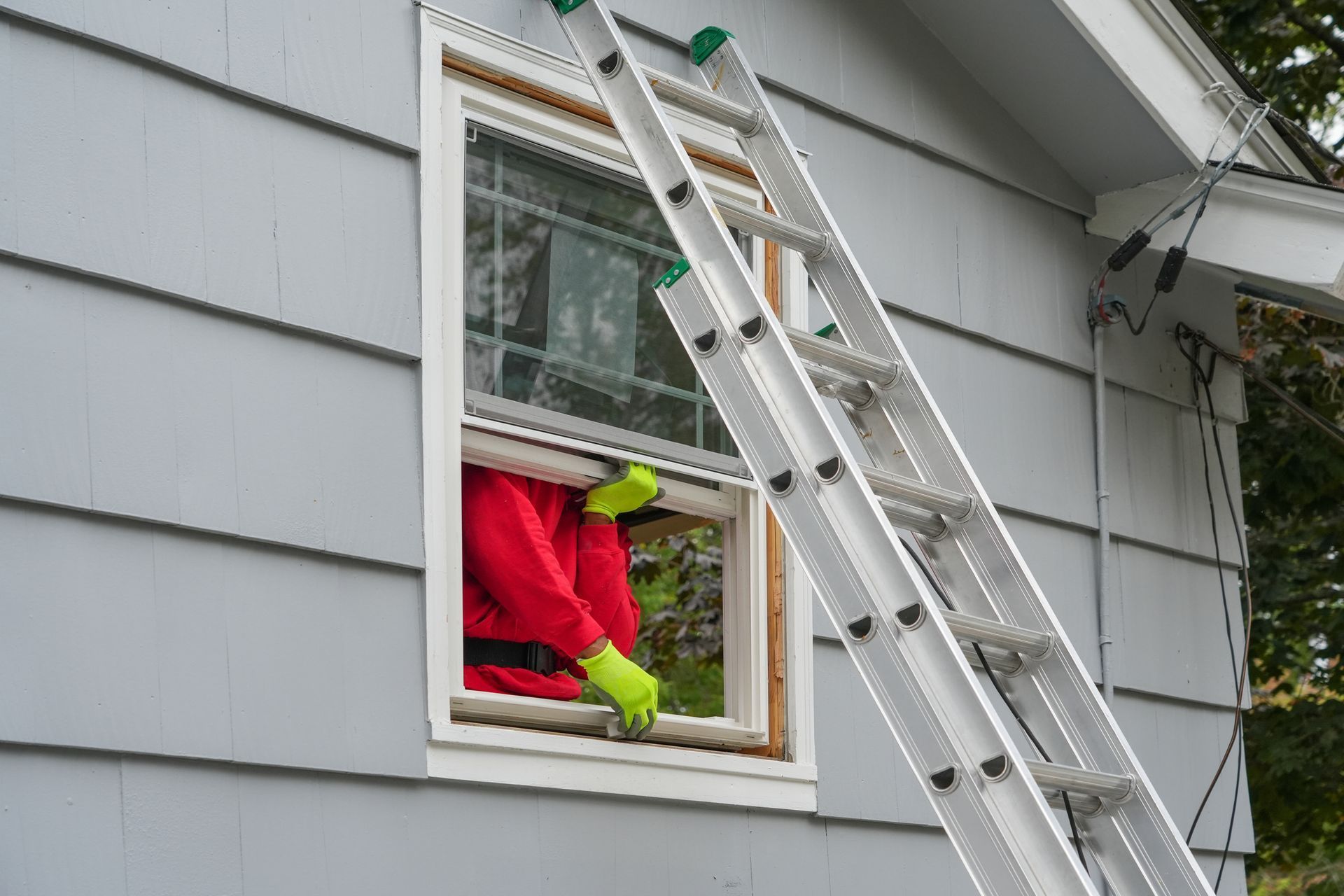 Person in red shirt and green gloves working on a window from a ladder outside a gray house. Person in red shirt and green gloves working on a window from a ladder outside a gray house.