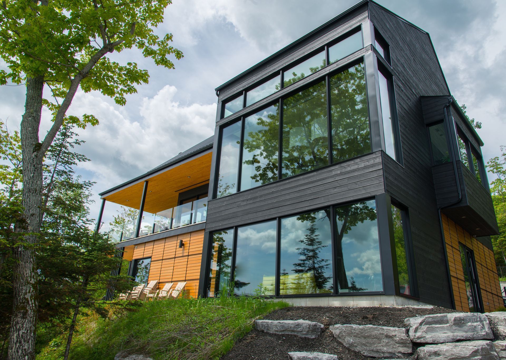 Modern black and wood home with large windows, covered patio, on a hillside with trees and cloudy sky.