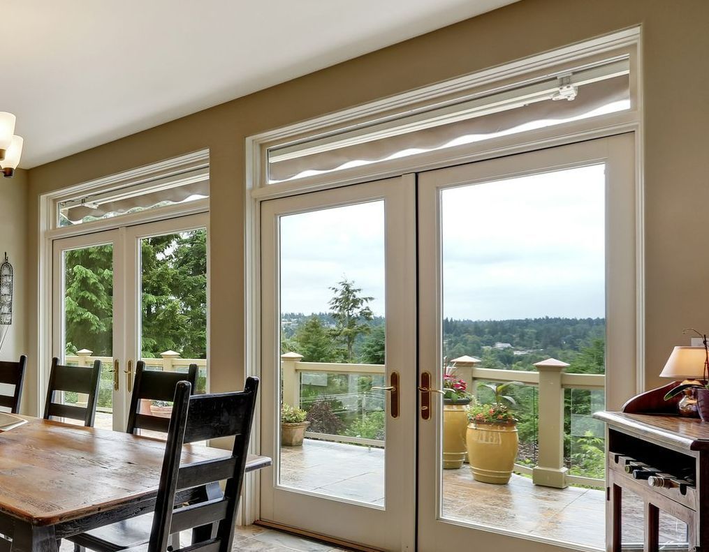 Dining room with glass doors leading to a balcony with trees in the distance; beige walls, wooden table and chairs. Dining room with glass doors leading to a balcony with trees in the distance; beige walls, wooden table and chairs.