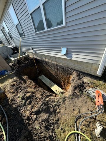A deep hole dug next to a house's foundation, with a wooden plank inside. Construction tools are nearby.