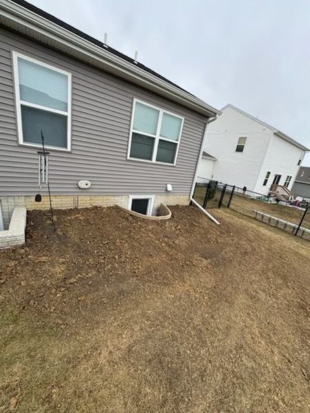 Side of a house with gray siding, dirt yard, window well, and black fence.