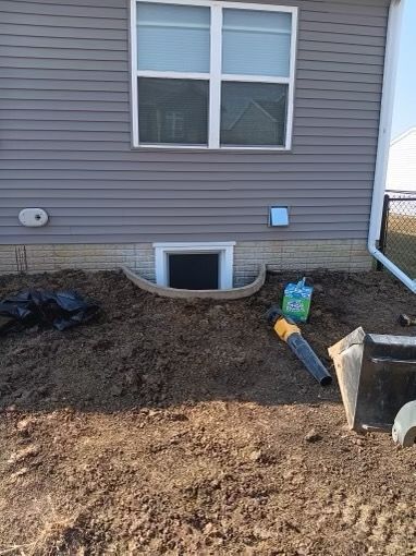 Exterior view of a house with a window well. Dirt, a leaf blower, and a small bag are present.