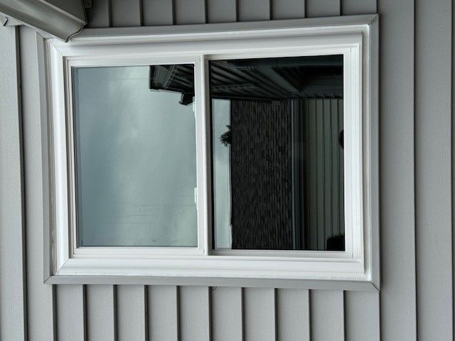 White-framed sliding window on gray siding, reflecting sky and interior of a building.