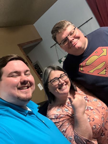 Three smiling people indoors, close up. One wears a blue polo, another a Superman shirt, and the third a floral top.