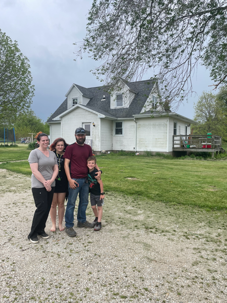 Family of four stands in front of a white house with a green yard on a cloudy day.