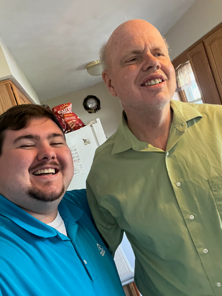 Two smiling people, one in a blue polo, one in a green shirt, indoors.
