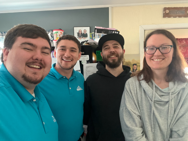 Four smiling people pose for a selfie indoors, two in blue shirts, one in black, and one in gray.