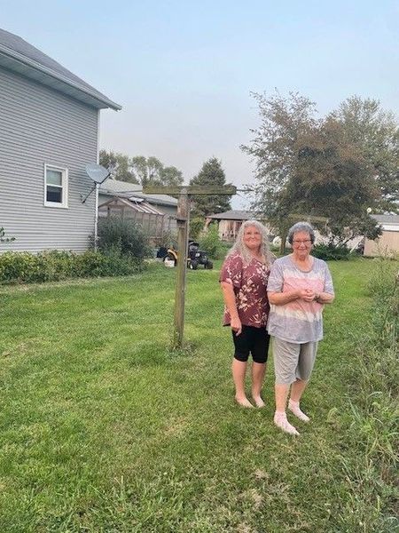 Two women standing barefoot in a grassy backyard, near a clothesline and a house.