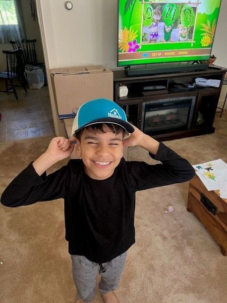 Boy wearing a blue and white cap, smiling, and holding his ears. TV shows a cartoon. Indoors.