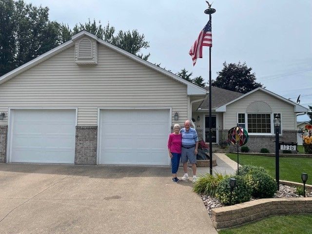 Couple standing in front of a house with an American flag. The house has a two-car garage and a well-kept lawn.
