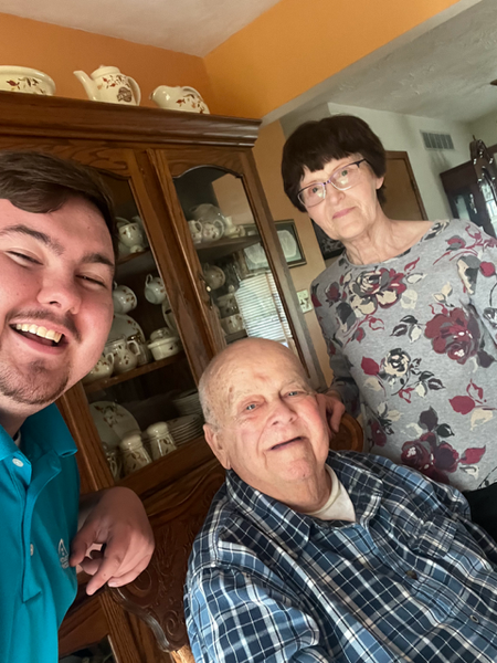 Man taking selfie with elderly man and woman; all smiling near china cabinet.