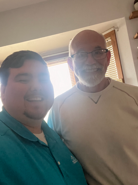 Two smiling people taking a selfie; one in a teal shirt, the other in a white shirt and glasses.