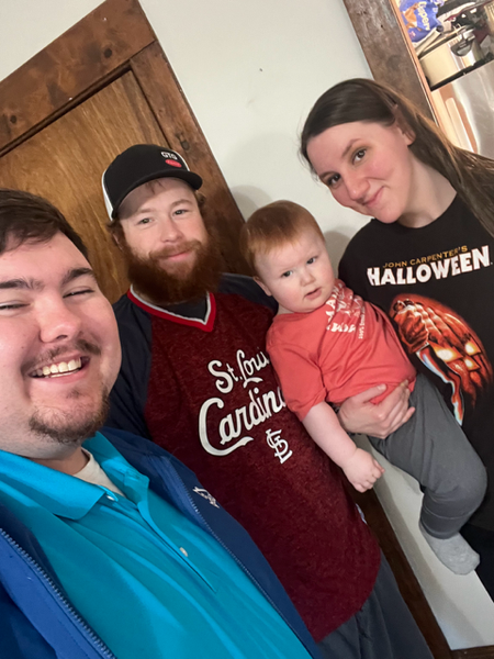 Four people smile for a selfie indoors: a man, a man with a beard, a woman holding a baby.