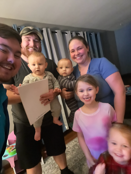Family of six, including two babies held by a man, smiling indoors. Another child and woman smile.