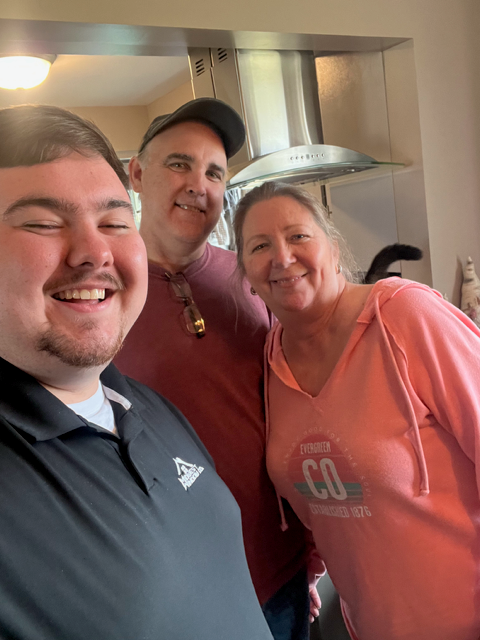 Three people smiling in a kitchen; a young man in a black shirt takes the selfie with a man in a red shirt and a woman in a pink top.