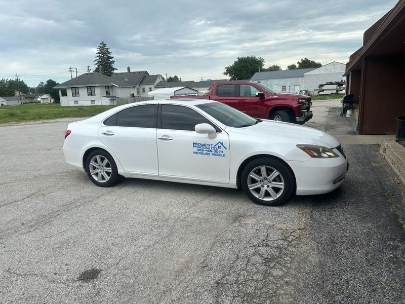 White sedan parked next to a building on a cloudy day; a red truck is also visible.