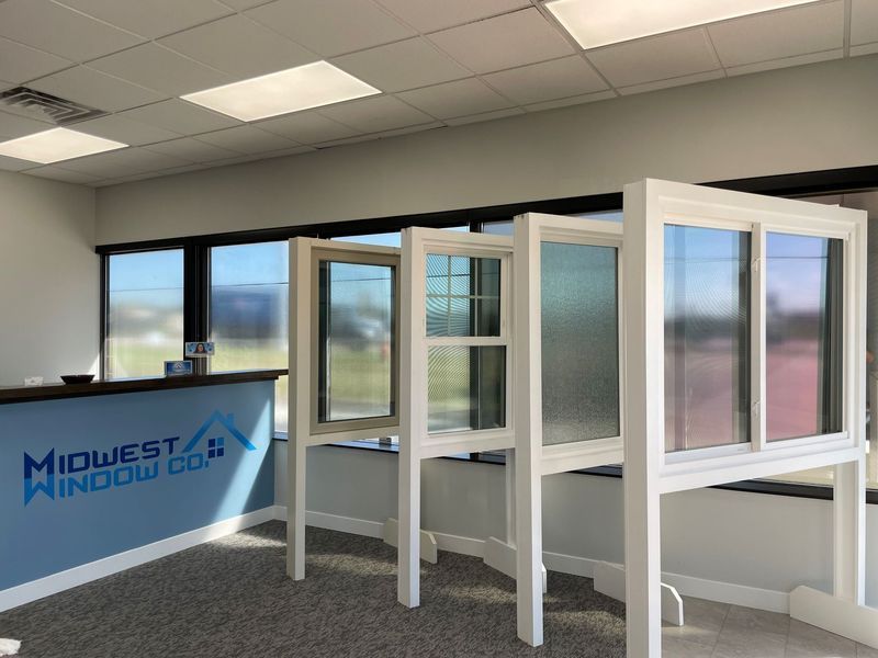 Interior of Midwest Window Co. showroom with window displays. Blue counter with logo, bright windows, and gray carpet.