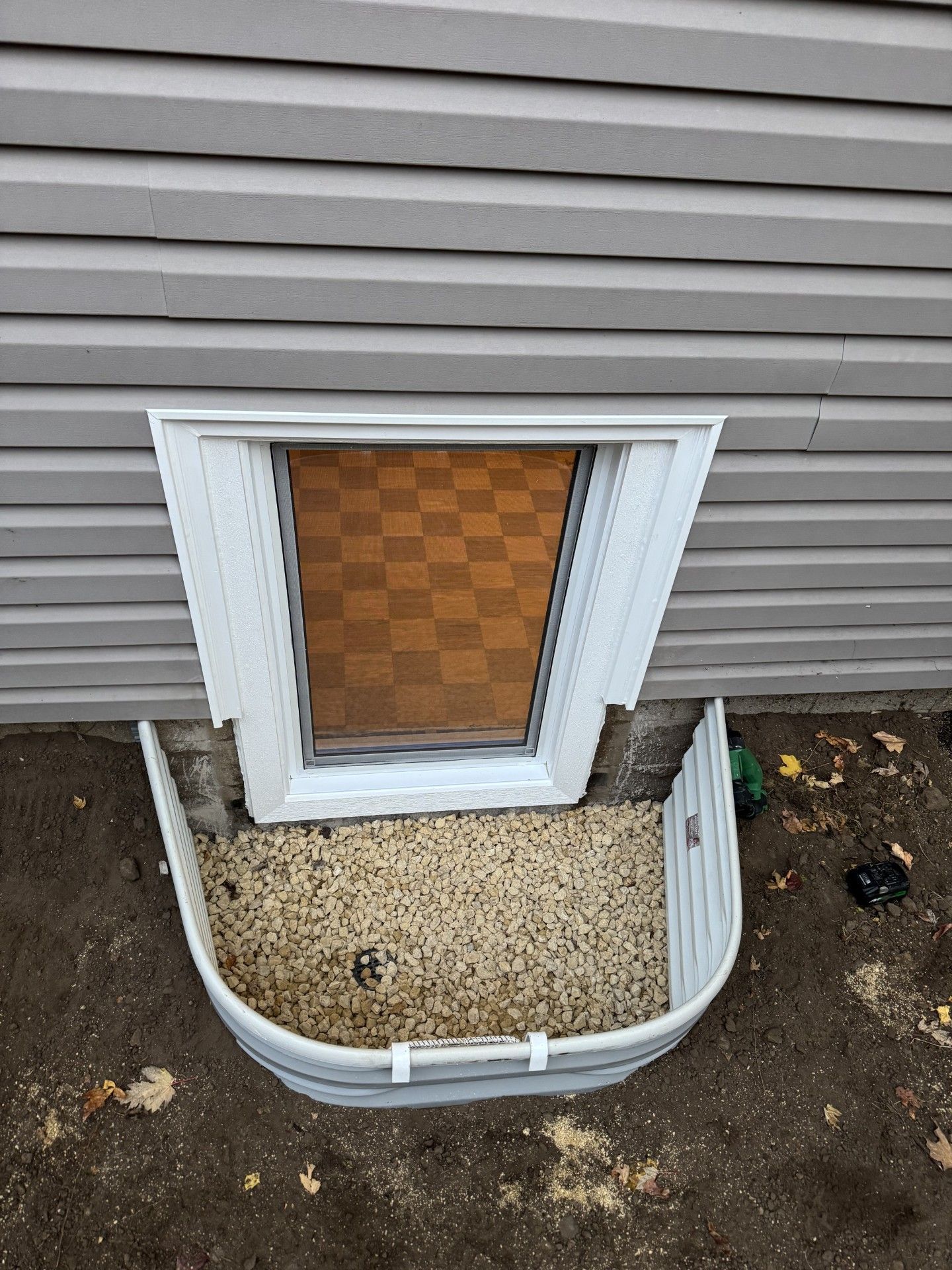 Window well with gravel, below a window with white trim, set in gray siding.