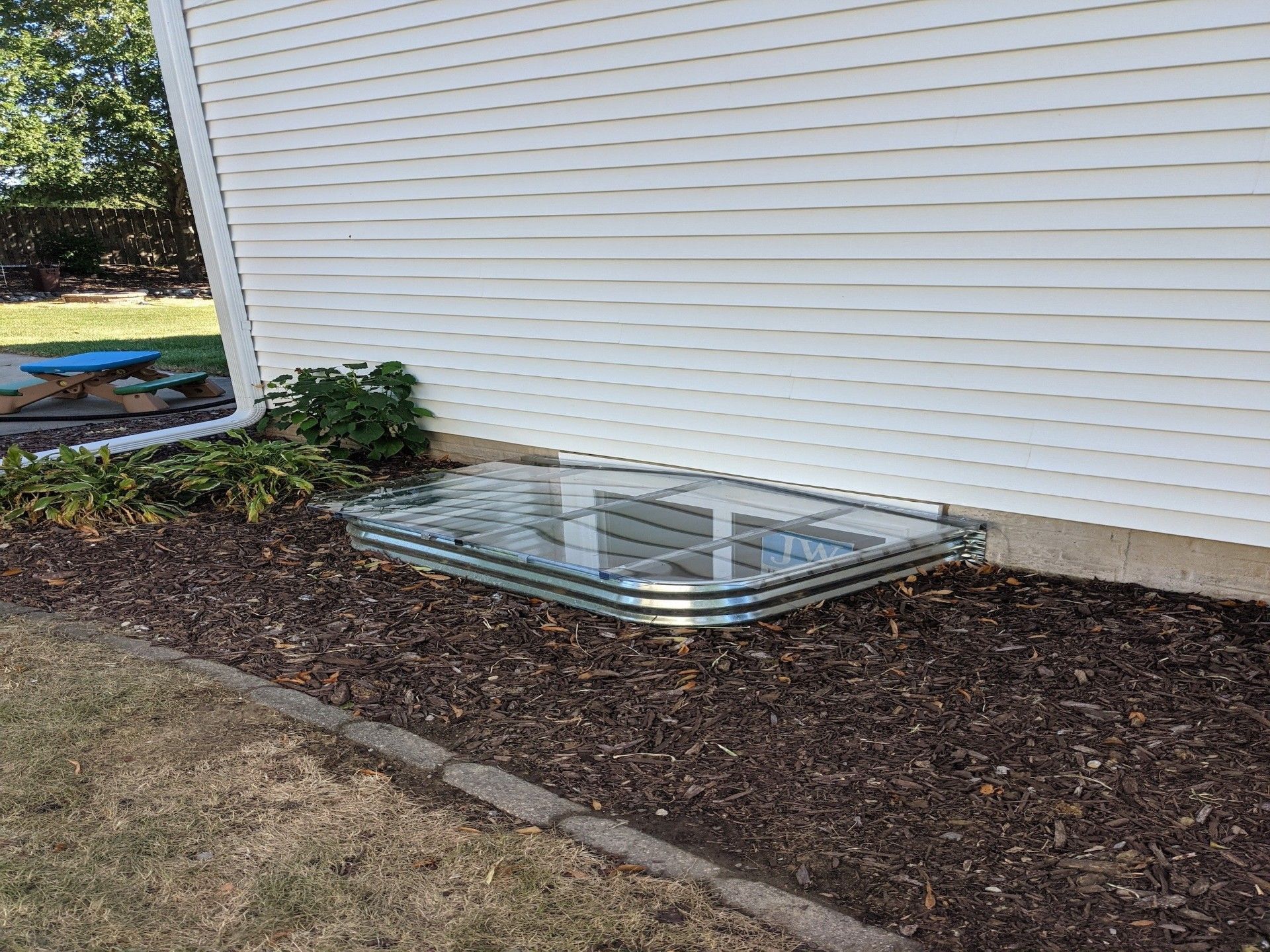 A window well with a clear cover is surrounded by mulch and the exterior wall of a white house.