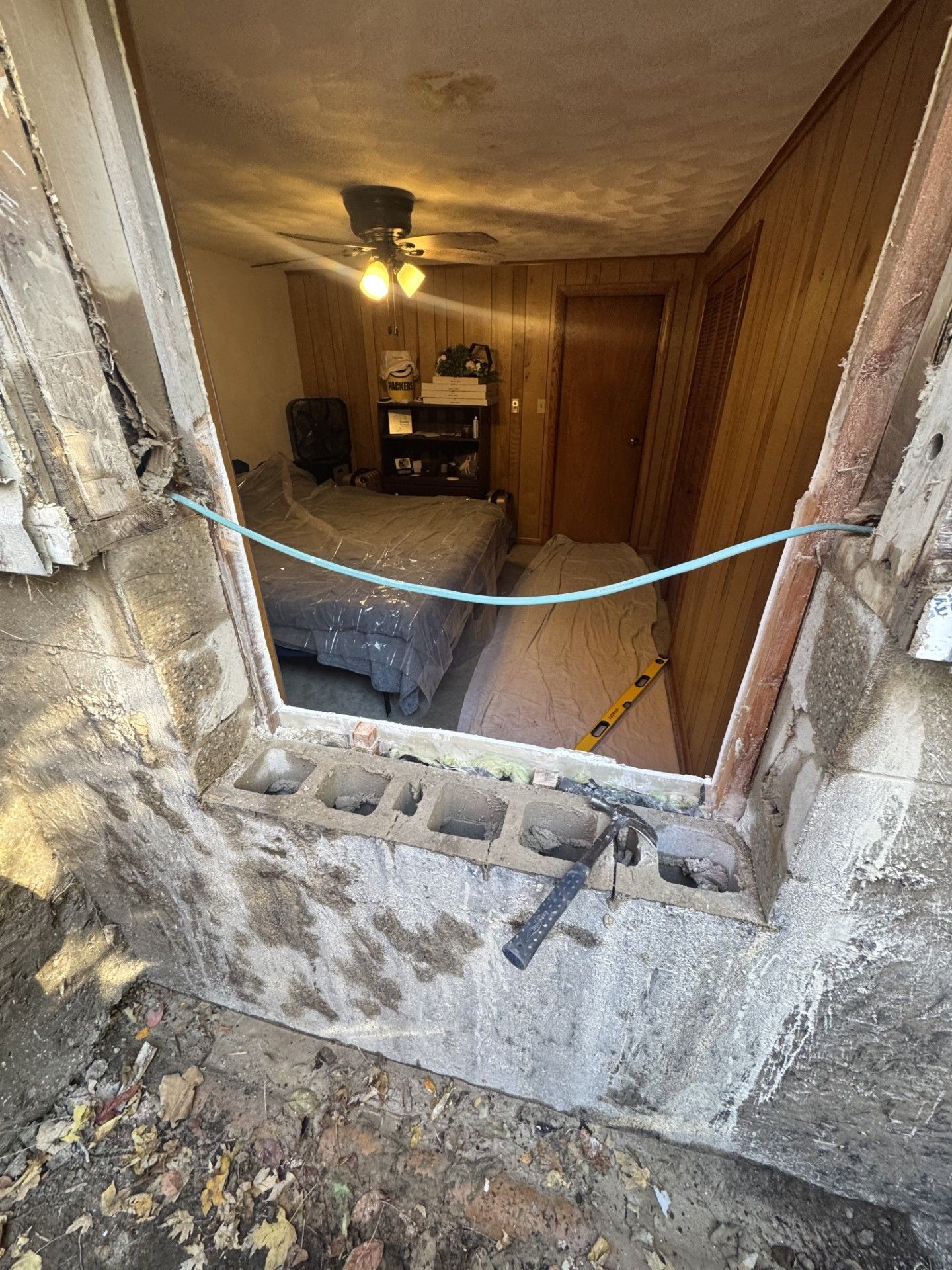View from a partially demolished opening into a room with a bed, dresser, and wooden paneling.