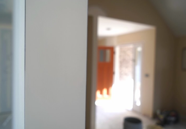 An extreme close-up of the edge of a wall with a smooth drywall surface. An open door is in the back