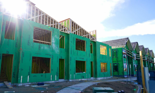 A row of green-wrapped, two-story townhouse buildings under construction with empty windows, wood fr