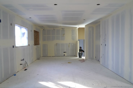 An unfinished room with drywall seams, ceiling cutouts, wiring, a window, and a person working near the far wall.