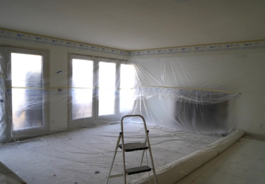 An empty room with covered windows and plastic sheeting over the floor and walls. A step ladder is in the middle of the room.