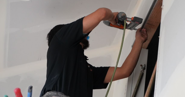 An installer is using a pole-mounted drywall sander with a vacuum hose along a white ceiling beside